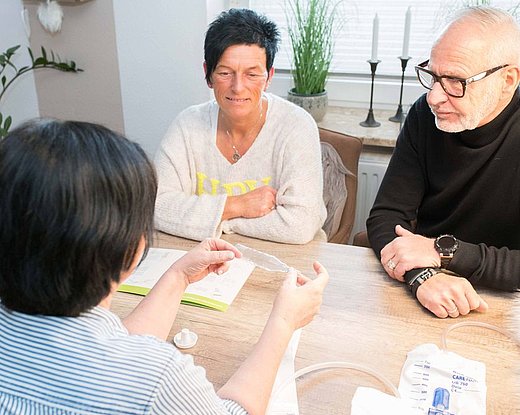 Eine Frau und ein Mann sitzen an einem Tisch gegenüber einer medizinischen Fachkraft, die ihnen ein medizinisches Gerät und einige Dokumente zeigt. Sie scheinen ein Gespräch in einem hellen, gemütlichen Raum zu führen.