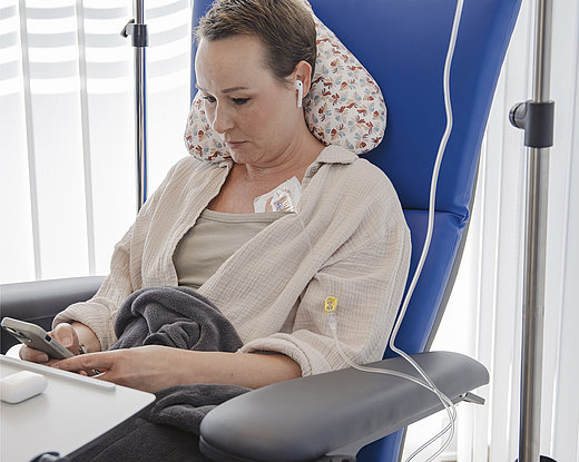 Frau sitzt in einem Therapiesessel mit Infusionsleitung am Arm, Ohrhörern im Ohr und Smartphone in der Hand; IV-Ständer und Decke sichtbar.
