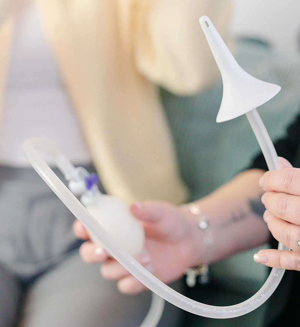 Nahaufnahme zweier Personen auf einem Sofa: Eine Patientin hält ein Darm-Management-/Irrigationssystem mit Schlauch, Trichter und Handpumpe in den Händen, während ihr gegenüber eine MPF-Mitarbeiterin die Anwendung erklärt.