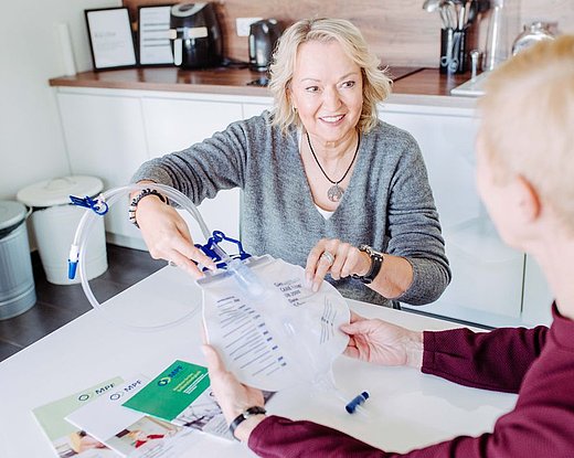 Beraterin zeigt einer Patientin am Tisch die Handhabung eines Urinbeutels mit Katheterschlauch; beide halten den Beutel, daneben liegen Unterlagen.