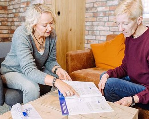 Zwei ältere Erwachsene sitzen an einem Tisch und gehen gemeinsam Dokumente durch. Eine Person zeigt auf die Papiere, während die andere aufmerksam zuhört. Beide scheinen sich in einem gemütlichen Raum mit einer Backsteinwand und einem orangefarbenen Sofa im Hintergrund zu unterhalten.