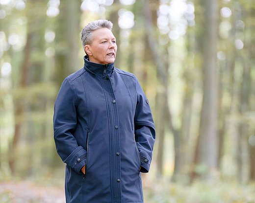 Eine Frau spaziert durch einen herbstlichen Wald. Sie trägt eine warme Jacke und wirkt ruhig und entspannt, während sie zwischen den gefallenen Blättern steht.