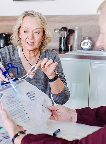 Zwei Frauen sitzen an einem Tisch und setzen einen medizinischen Urinsammelbeutel mit Schläuchen zusammen. Eine Frau demonstriert den Aufbau, während die andere den Beutel hält. Auf dem Tisch liegen medizinische Broschüren. Der Schauplatz scheint eine Küche zu sein.