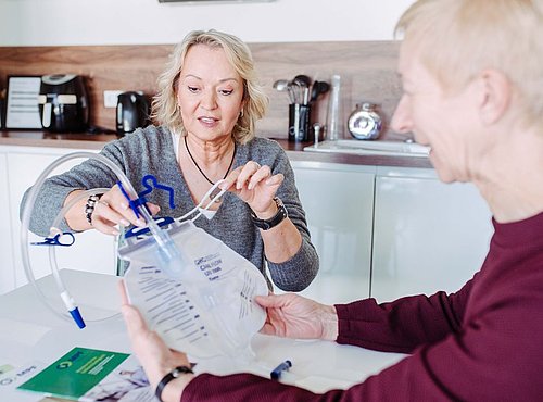 Zwei Frauen sitzen an einem Tisch und setzen einen medizinischen Urinsammelbeutel mit Schläuchen zusammen. Eine Frau demonstriert den Aufbau, während die andere den Beutel hält. Auf dem Tisch liegen medizinische Broschüren. Der Schauplatz scheint eine Küche zu sein.