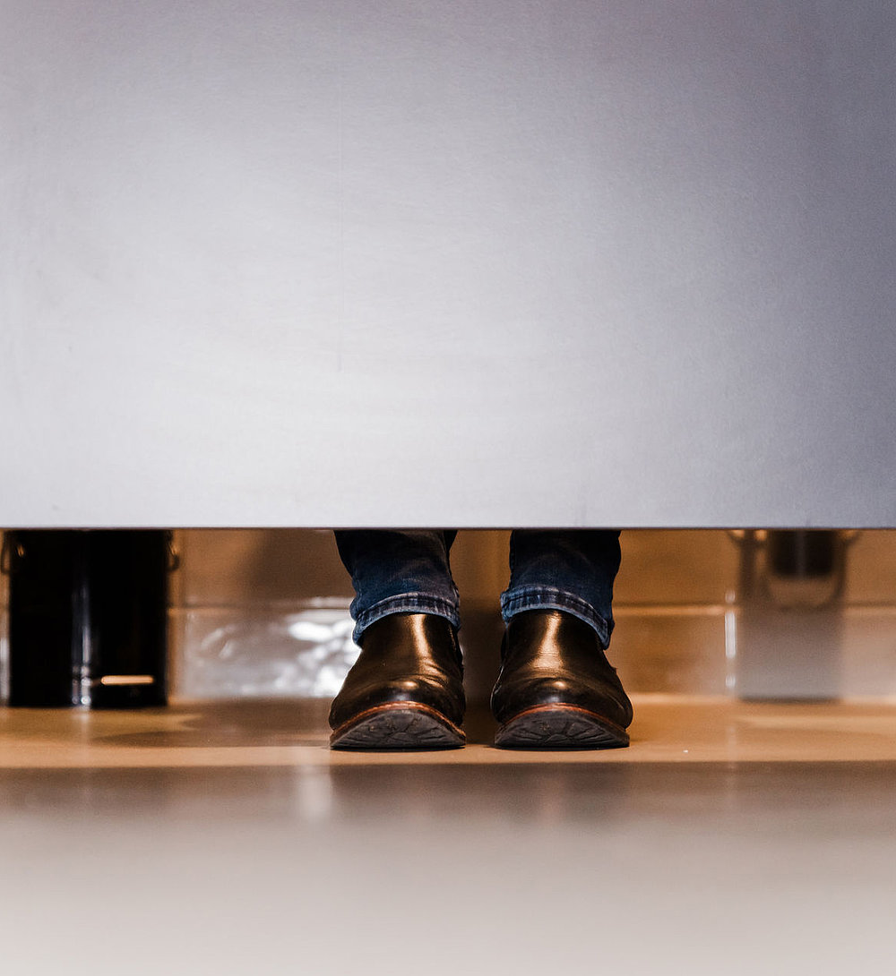Blick unter einer geschlossenen Toilettenkabine: Unter der Tür sind die Hosenbeine und braunen Lederschuhe einer Person zu sehen