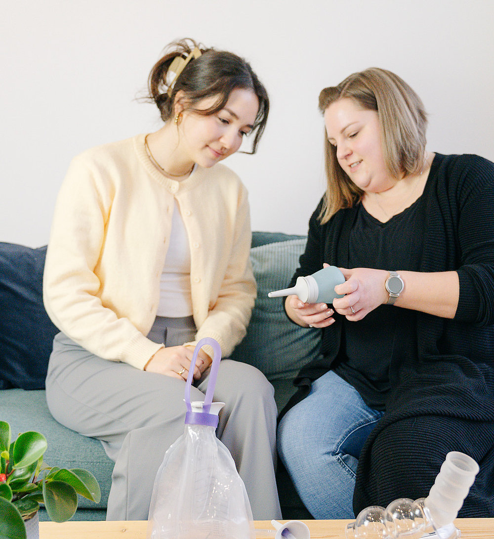 Zwei Frauen sitzen auf einem Sofa; eine Patientin erklärt der anderen die Handhabung einer kleinen Irrigationspumpe, auf dem Couchtisch davor liegen weitere Darm-Management-Hilfsmittel und ein Demonstrationsmodell.